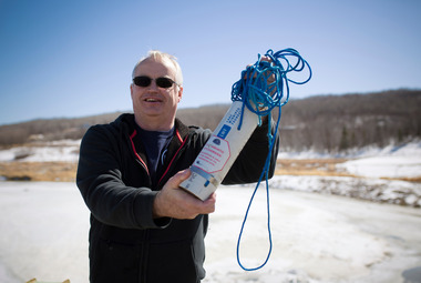 A middle-age white man wearing sunglasses holds an integrated water sampler towards the camera, with a winter landscape in the background.