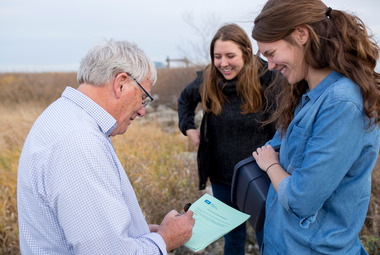 An older man and two younger women standing in a field, looking at a data-collection form