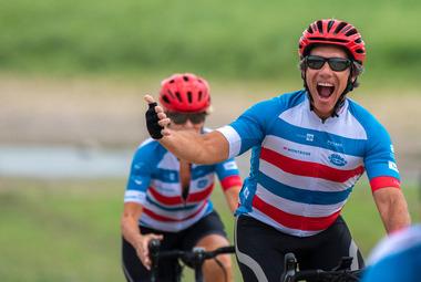 A man on a bike wearing a striped cycling jersey smiles and gestures towards the camera