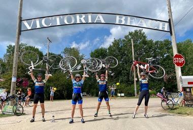 Four people wearing matching cycling gear standing underneath the Victoria Beach sign, holding their bikes over their heads.
